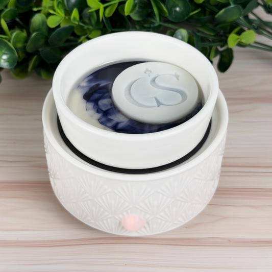 Embossed “GS” wax melt tart melting in a white electric wax warmer dish (blue-purple wax visible), styled on a light wood background.