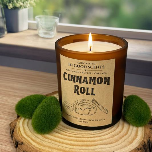 Lit Cinnamon Roll 8oz amber jar candle by In Good Scents on a wood slice pedestal near a window, surrounded by green moss accents.