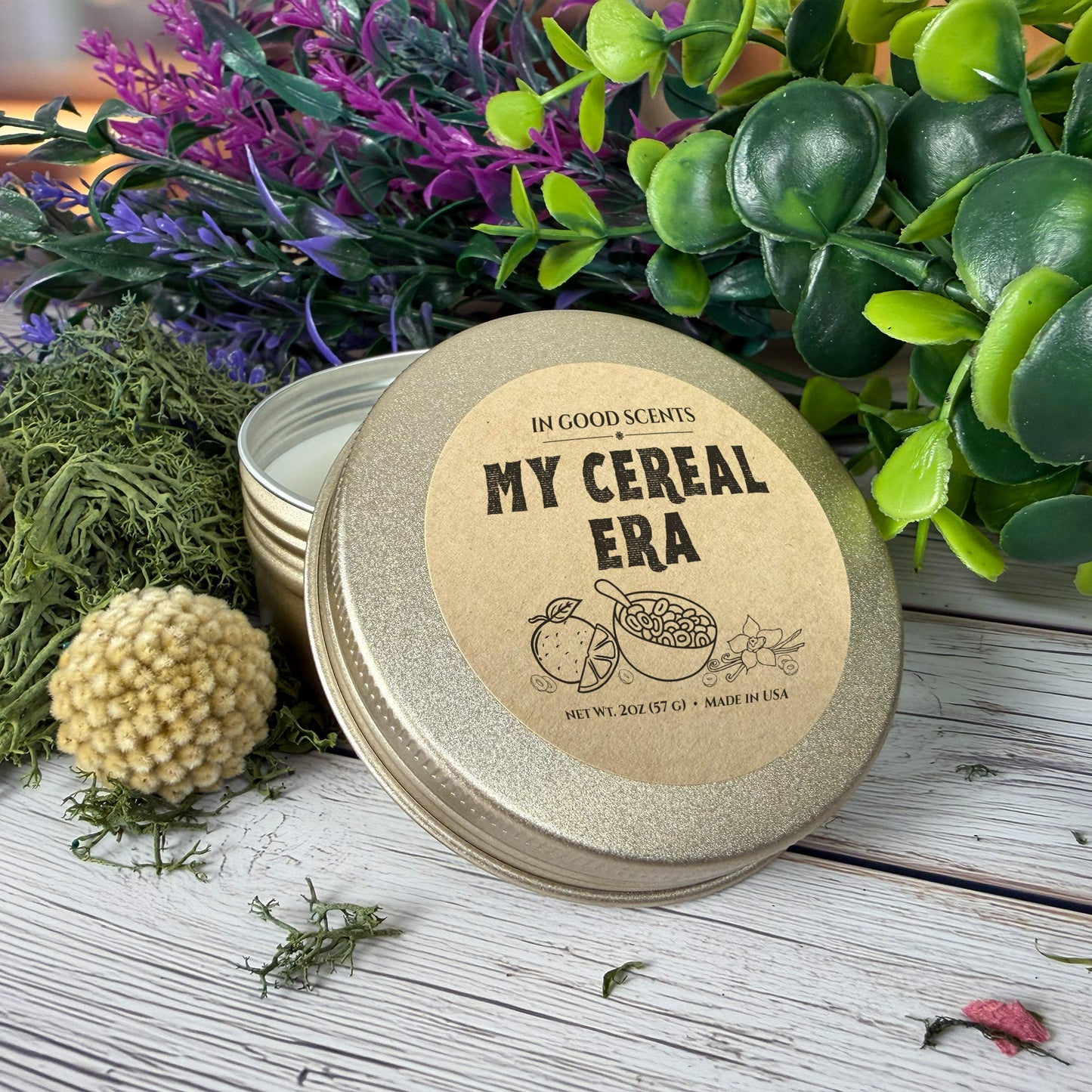My Cereal Era 2oz travel tin candle by In Good Scents, tin open with lid leaning forward, styled on white wood with moss and greenery.