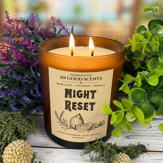 Lit Night Reset 12oz candle (In Good Scents) showing two flames in an amber jar, surrounded by purple florals, greenery, and moss on a rustic wood background.