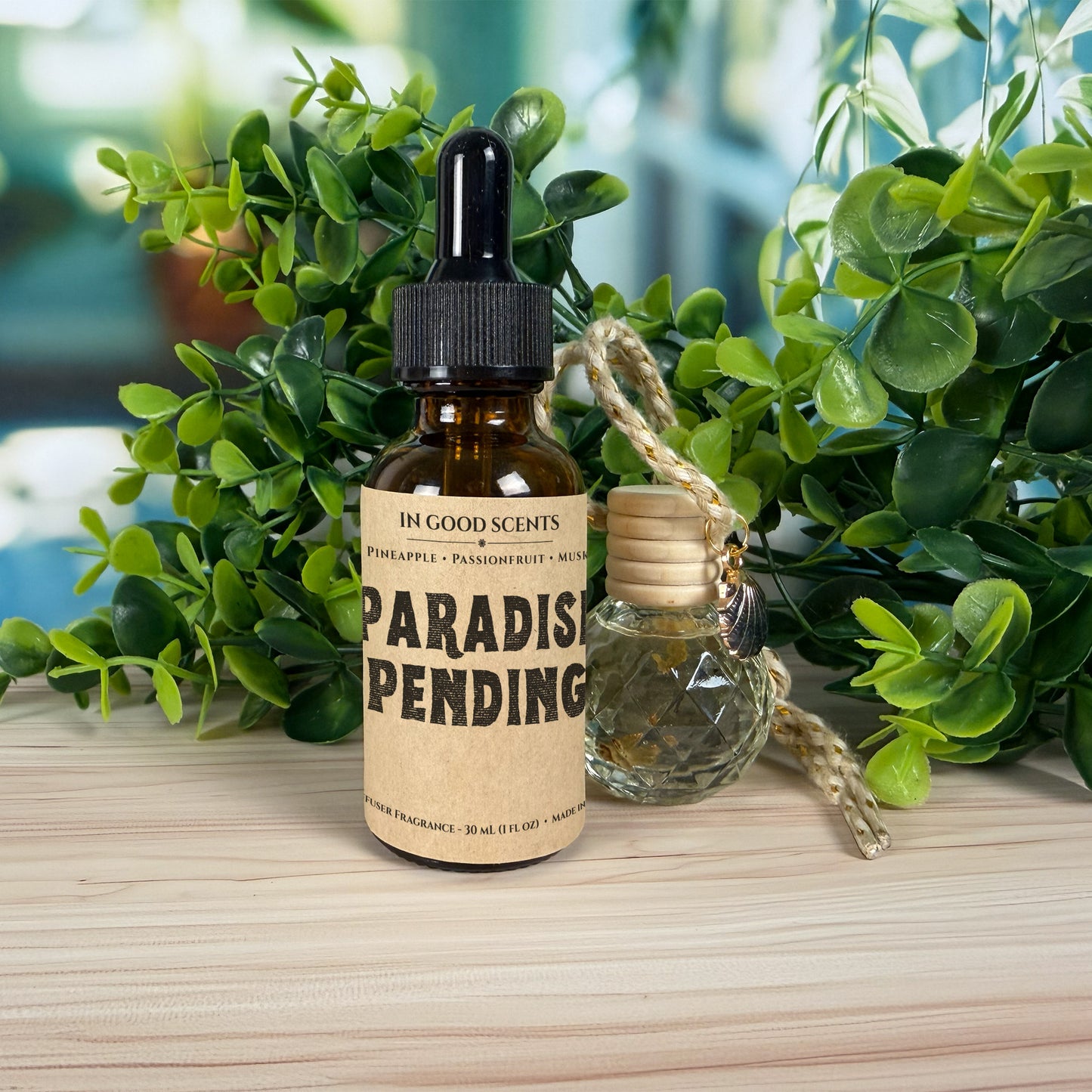 Bottle of 'Paradise Pending' with a dropper and hanging diffuser on a wooden surface with green plants in the background.