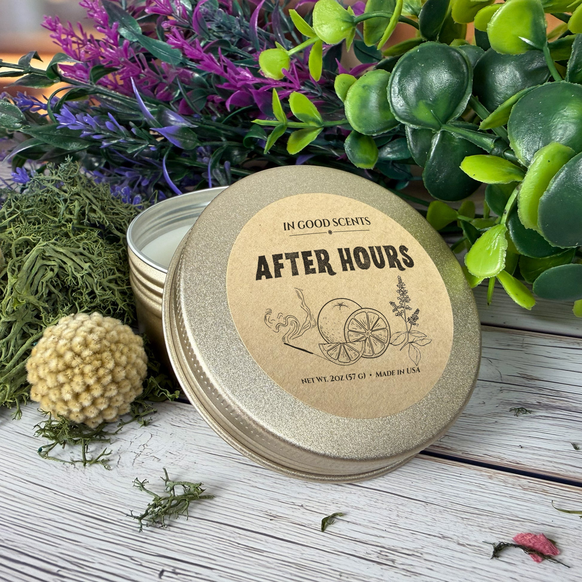 After Hours 2oz tin candle by In Good Scents, close-up top label shot on a rustic tabletop with greenery and neutral decor styling.