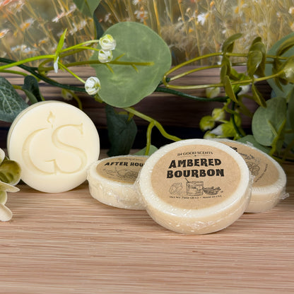 Group of In Good Scents wrapped wax melt tarts, featuring Ambered Bourbon .75 oz in front beside the GS logo wax melt on a wood tabletop with greenery.
