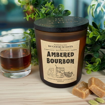 Ambered Bourbon 12oz candle in amber jar with black lid, beside a whiskey glass and butterscotch cubes, In Good Scents.