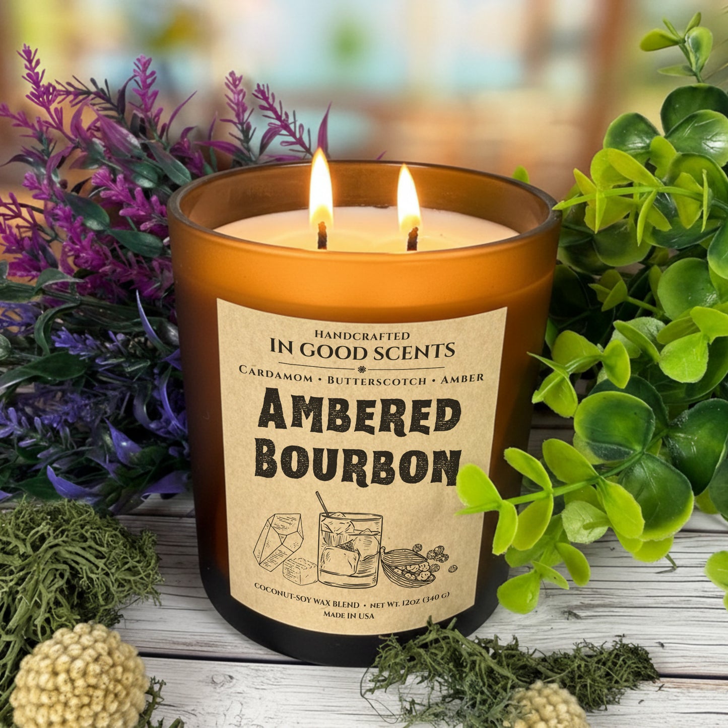 Lit Ambered Bourbon 12oz amber jar candle with two flames, styled with greenery and purple florals, In Good Scents.