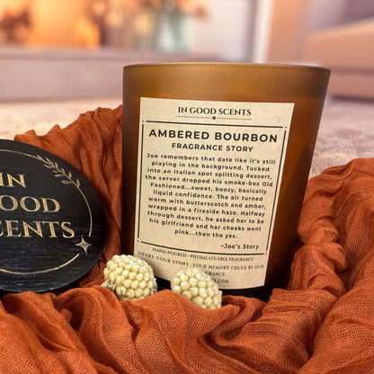Ambered Bourbon fragrance story label on 12oz amber jar candle styled on rust fabric with black In Good Scents lid.