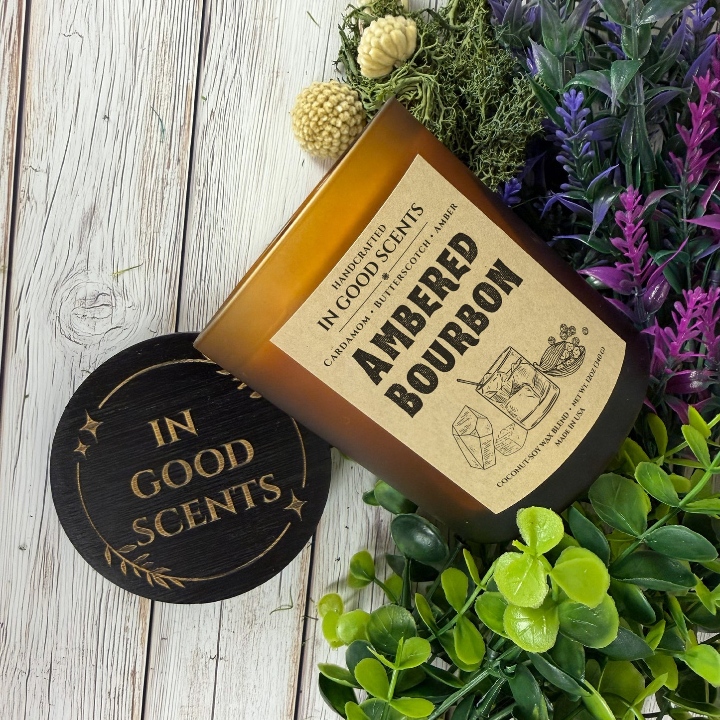 Ambered Bourbon 12oz candle laying diagonally with black In Good Scents lid, styled with greenery on white wood surface.