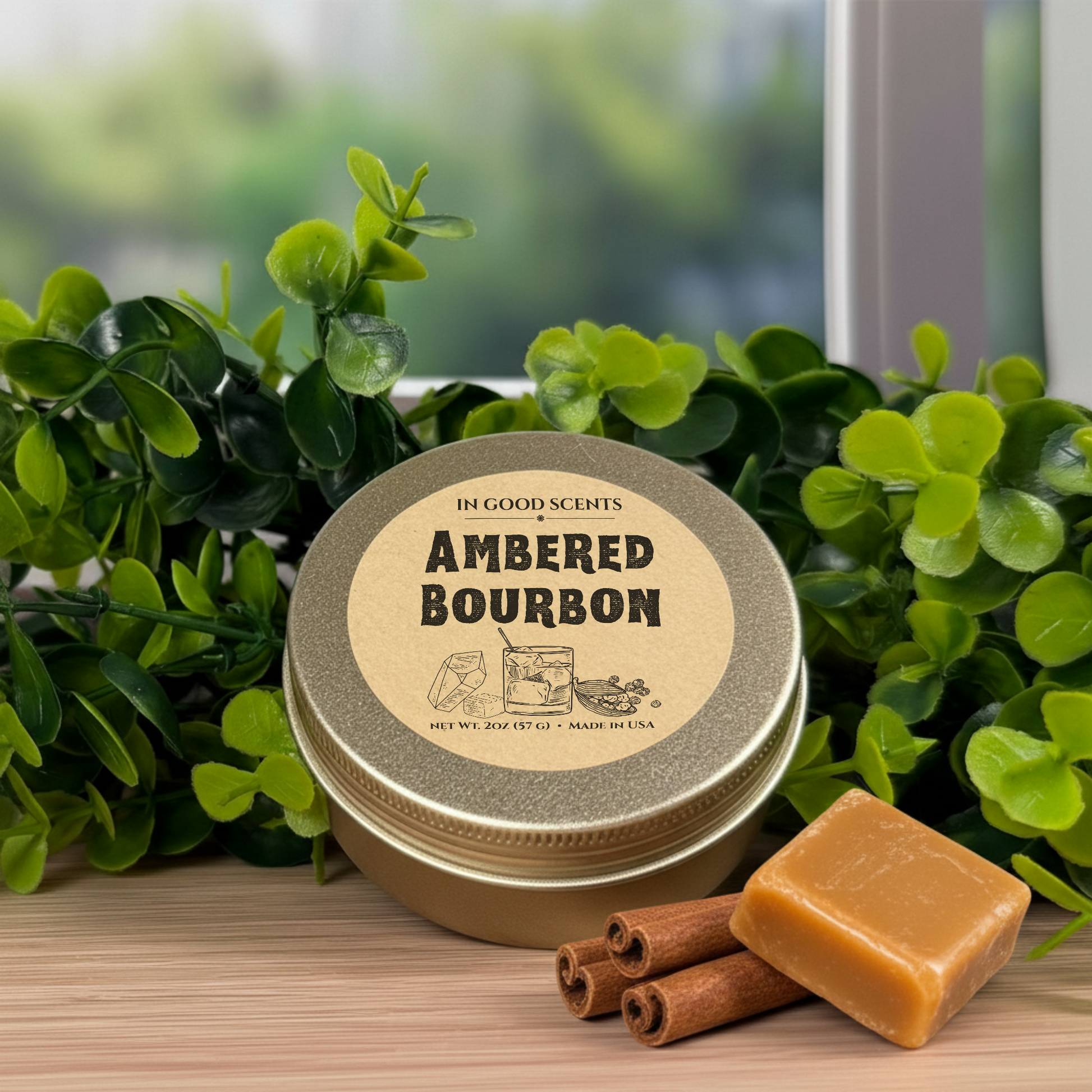 In Good Scents Ambered Bourbon 2oz travel tin candle with kraft lid label, styled with greenery, caramel cube, and cinnamon sticks on a wood surface.
