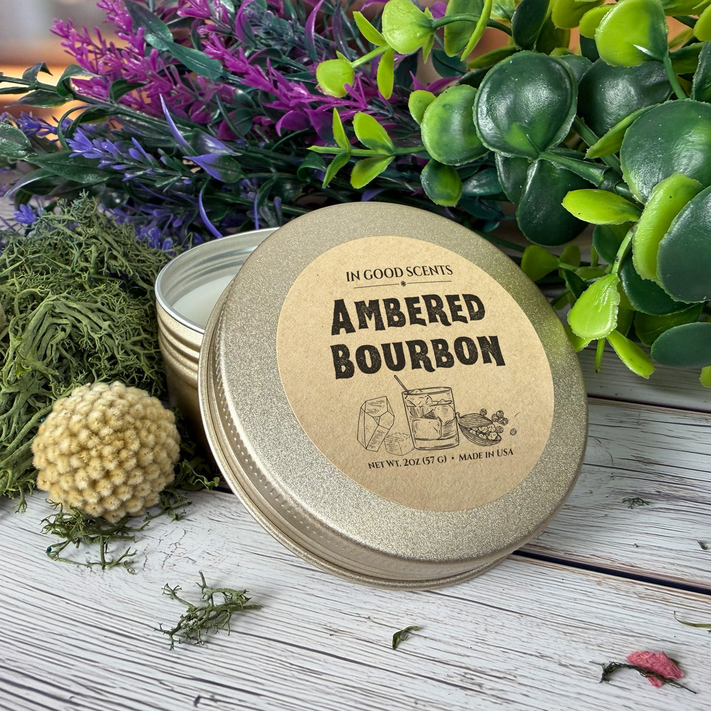 In Good Scents Ambered Bourbon 2oz travel tin candle close-up of the labeled lid on a rustic white wood surface with greenery and flowers.
