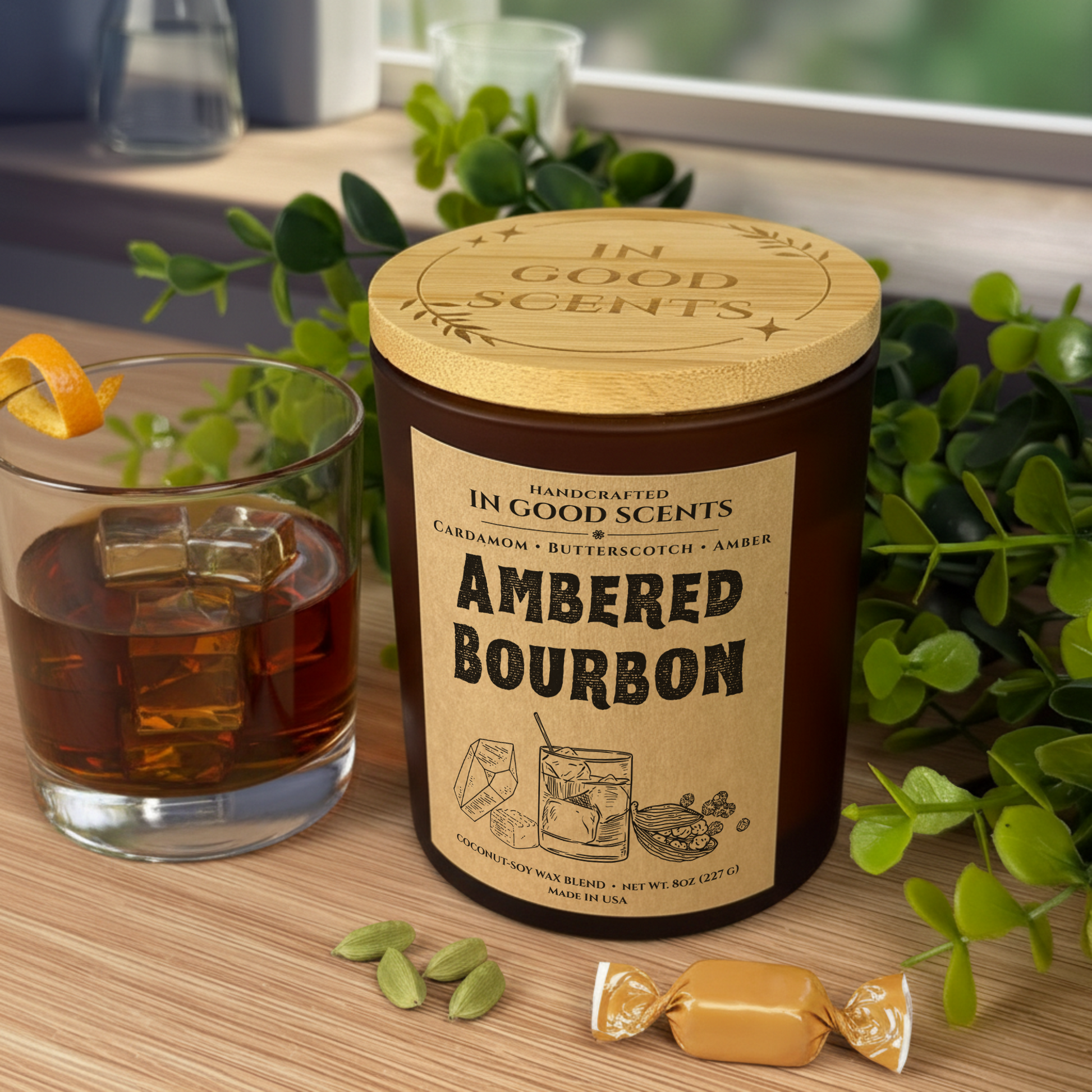 Ambered Bourbon 8oz candle with bamboo lid in an amber glass jar on a windowsill, styled beside a bourbon rocks glass with ice, cardamom pods, and a wrapped butterscotch candy.