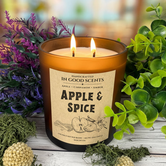 Apple & Spice candle lit — In Good Scents 12oz amber jar with two flames, kraft label, styled with greenery and purple florals on rustic wood.