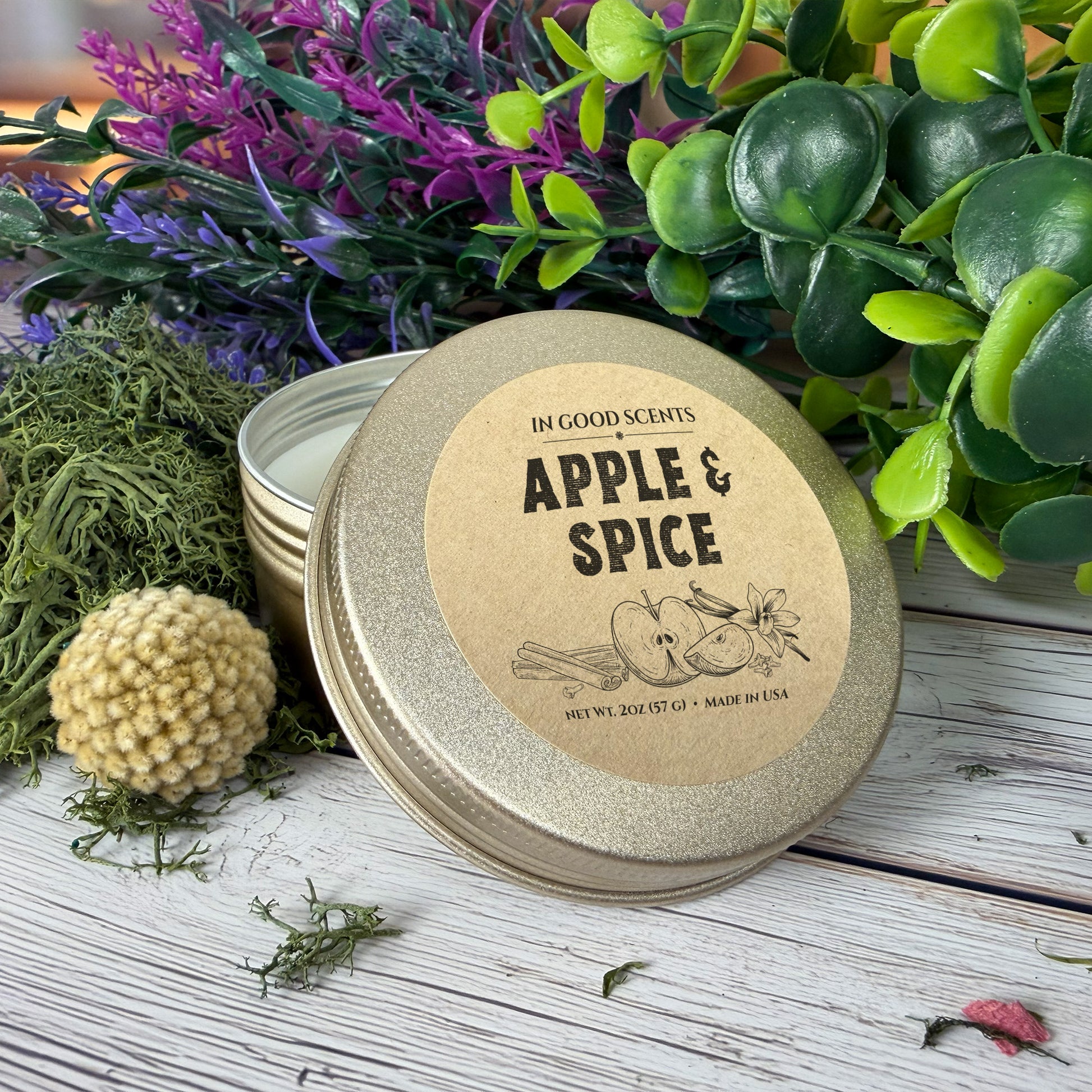 Apple & Spice 2oz candle tin with kraft lid label on a rustic wood background with greenery and moss accents.