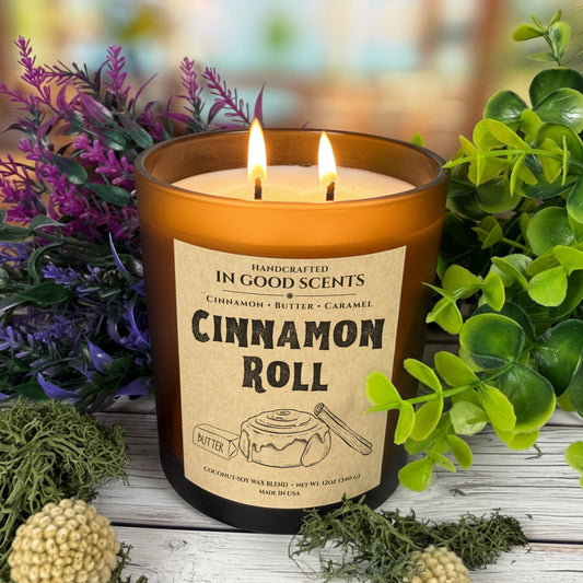 Cinnamon Roll 12oz amber jar candle lit with two flames, label facing front, surrounded by greenery and purple florals