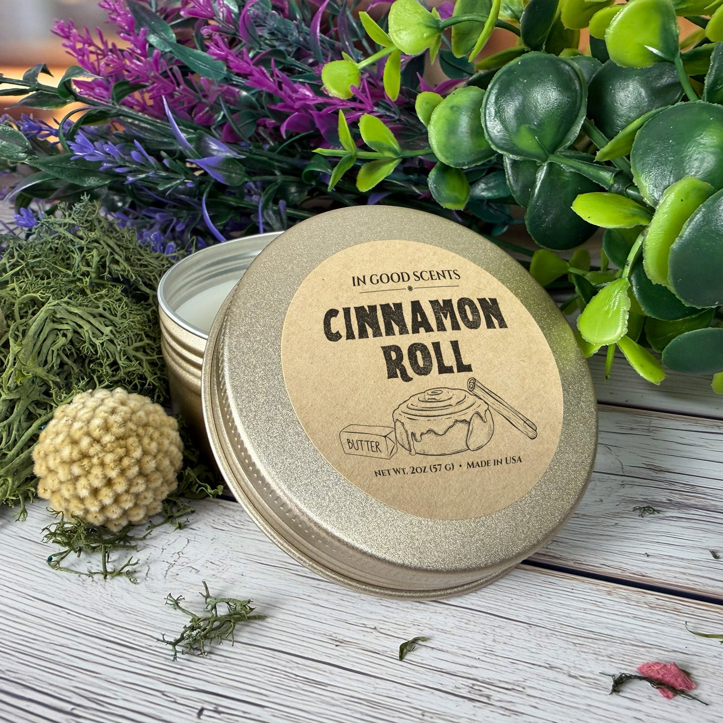 In Good Scents “Cinnamon Roll” 2oz travel tin candle with lid leaned forward on whitewashed wood with greenery and moss accents.