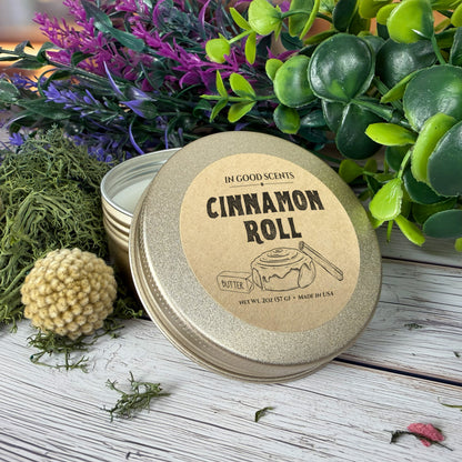 In Good Scents “Cinnamon Roll” 2oz travel tin candle with lid leaned forward on whitewashed wood with greenery and moss accents.