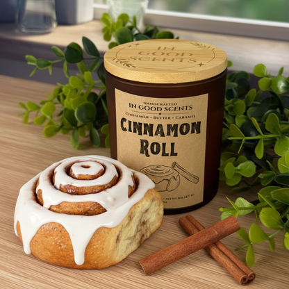 Cinnamon Roll 8oz candle with bamboo lid by In Good Scents, styled with an iced cinnamon roll and cinnamon sticks on a wooden table with greenery.