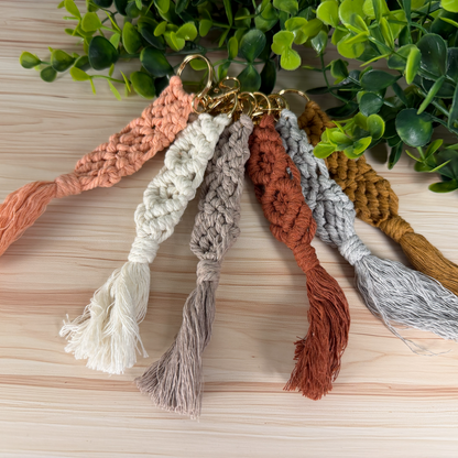 Set of macrame keychains in various colors on a wooden surface with green leaves in the background.