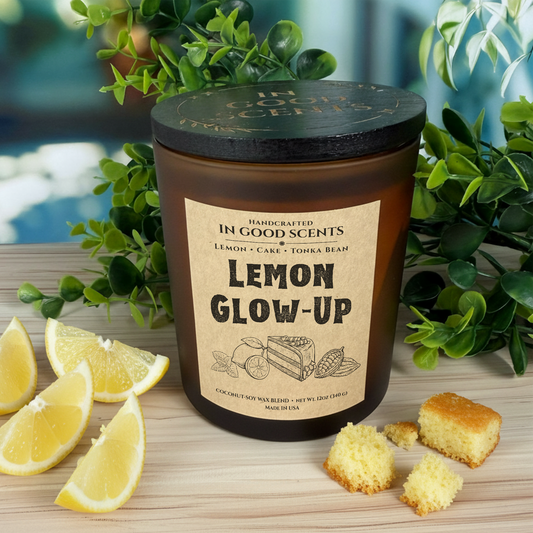 Lemon Glow-Up 12oz candle in amber jar with black lid, styled with lemon wedges and cake crumbs on wood surface.