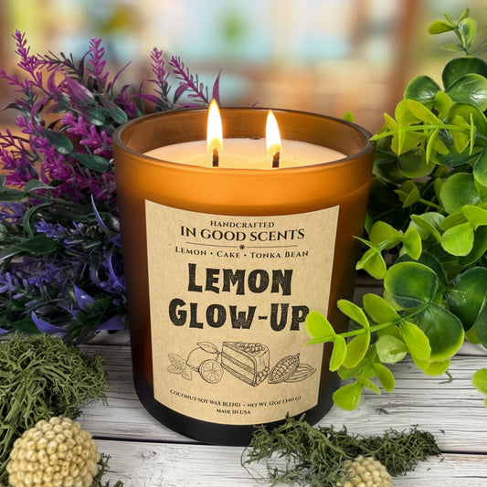 Lemon Glow-Up 12oz candle lit (double wick) in amber jar, surrounded by greenery and purple florals on rustic wood background.