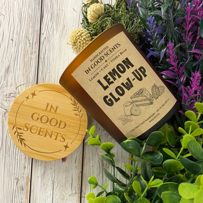 Lemon Glow-Up 8oz candle and bamboo lid styled on white wood surface with greenery and purple florals, overhead angle.