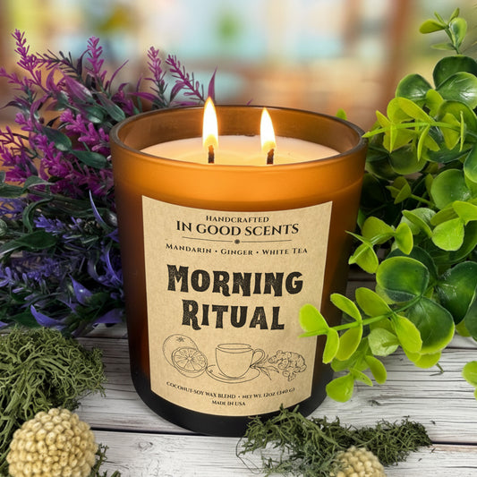 Lit Morning Ritual 12oz amber jar candle with two flames surrounded by greenery and purple florals
