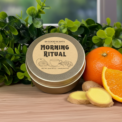 Morning Ritual 2oz travel tin candle by In Good Scents on a wood surface with greenery, fresh orange, and sliced ginger beside the labeled lid.