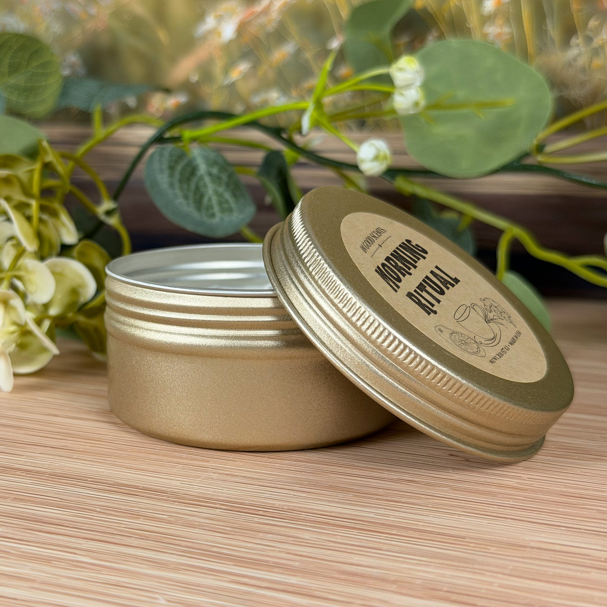 Morning Ritual 2oz travel tin candle by In Good Scents shown from the side with the lid leaning against the tin, surrounded by soft greenery on a wood surface.