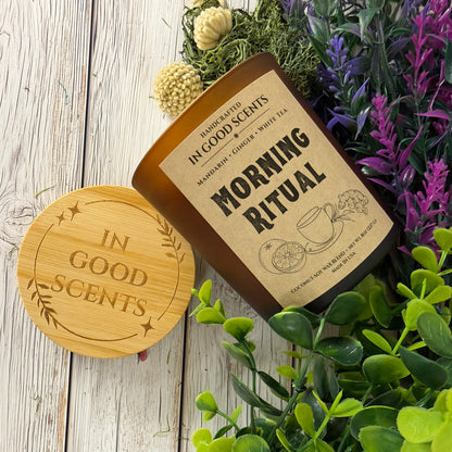 Morning Ritual 8oz amber jar candle with bamboo lid, angled top-down on white wood surface with greenery and purple florals.