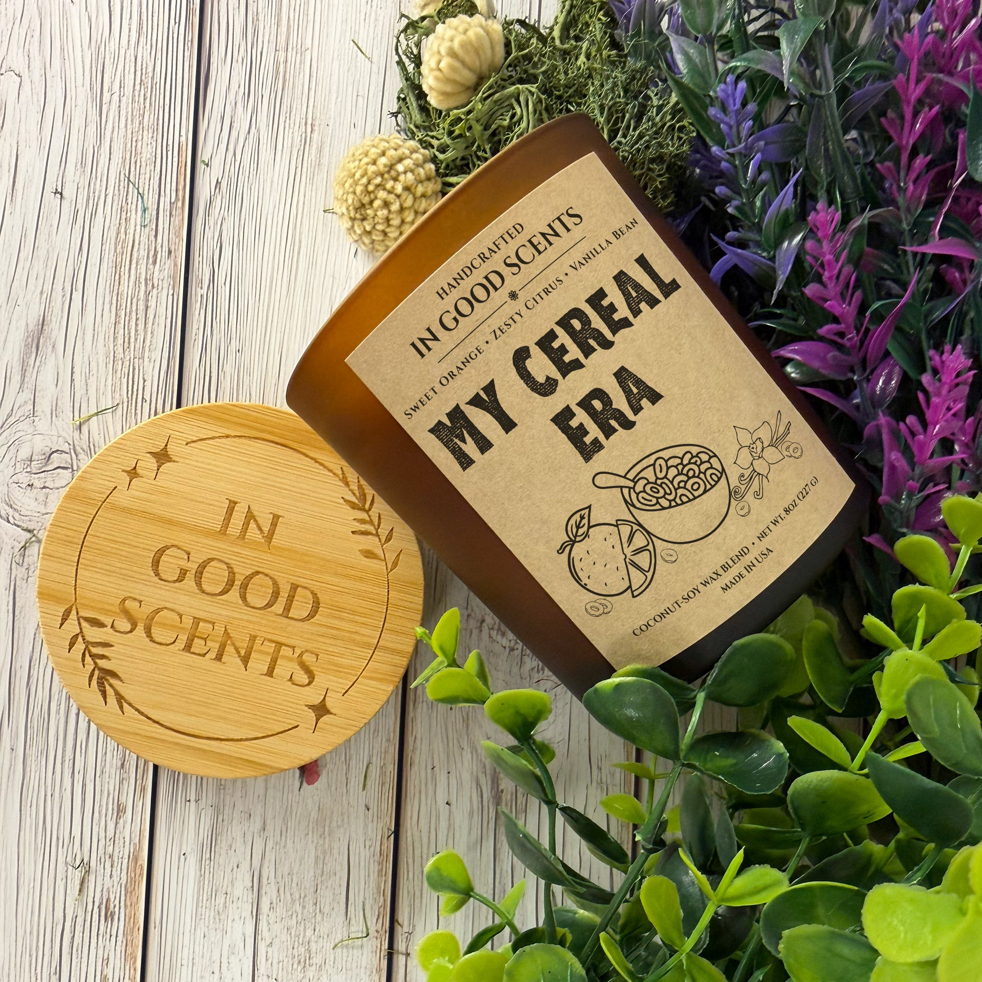 Flat lay of In Good Scents “My Cereal Era” 8oz candle with bamboo lid, surrounded by greenery on a rustic white wood surface.