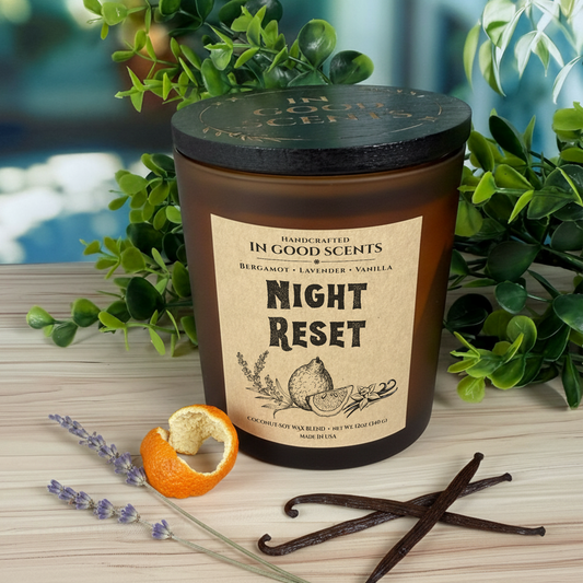 Night Reset 12oz candle (In Good Scents) in an amber jar with black lid, styled with greenery, lavender sprig, orange peel, and vanilla beans on a light wood surface.