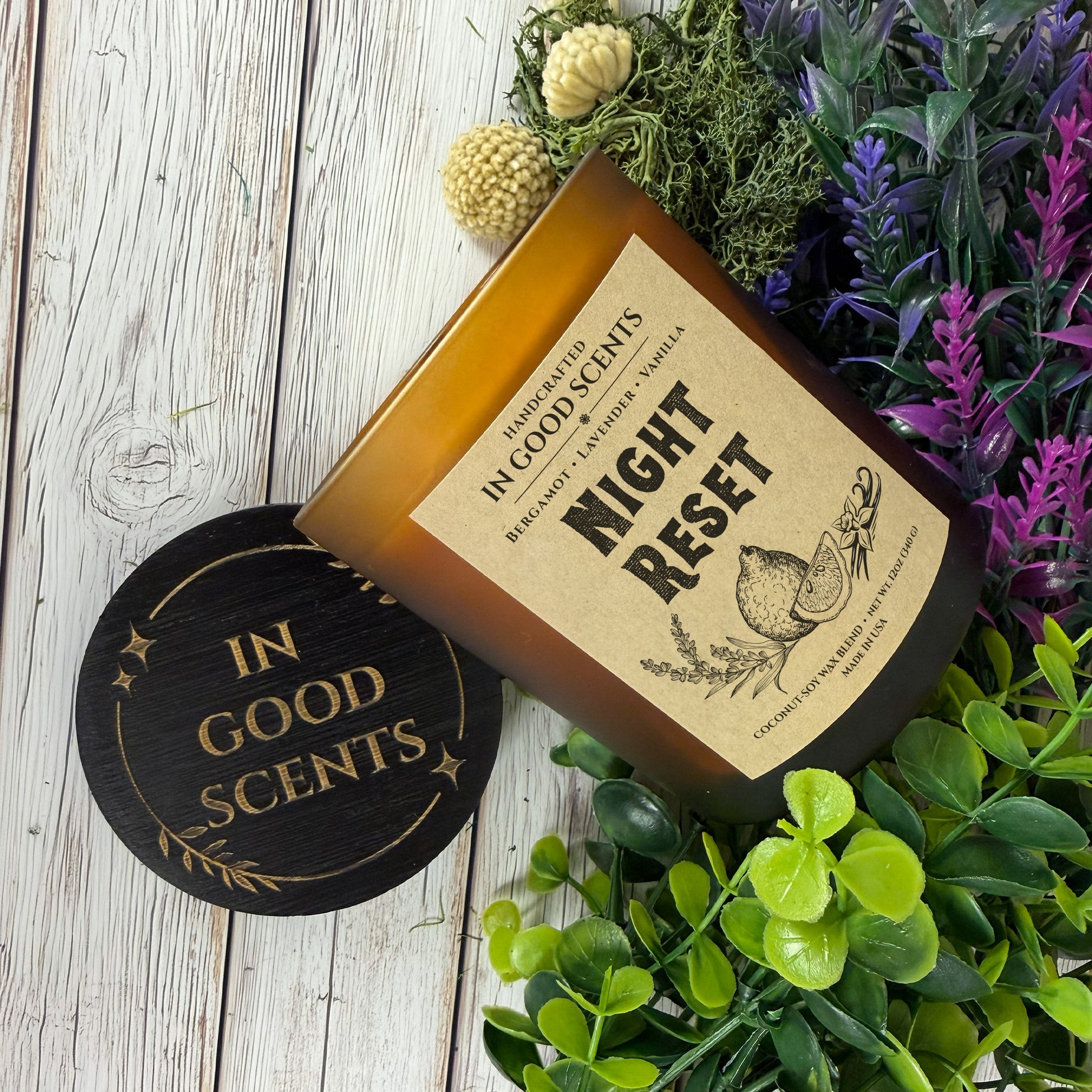 Amber jar “Night Reset” 12oz coconut-soy candle lying on its side beside black In Good Scents lid on white wood surface with greenery, moss, and purple florals.