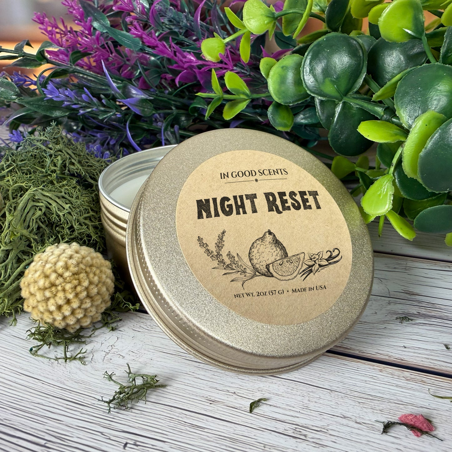 Night Reset 2oz Travel Tin Candle close-up with lid leaning against the tin, styled with greenery, purple florals, and moss on a rustic wood background.