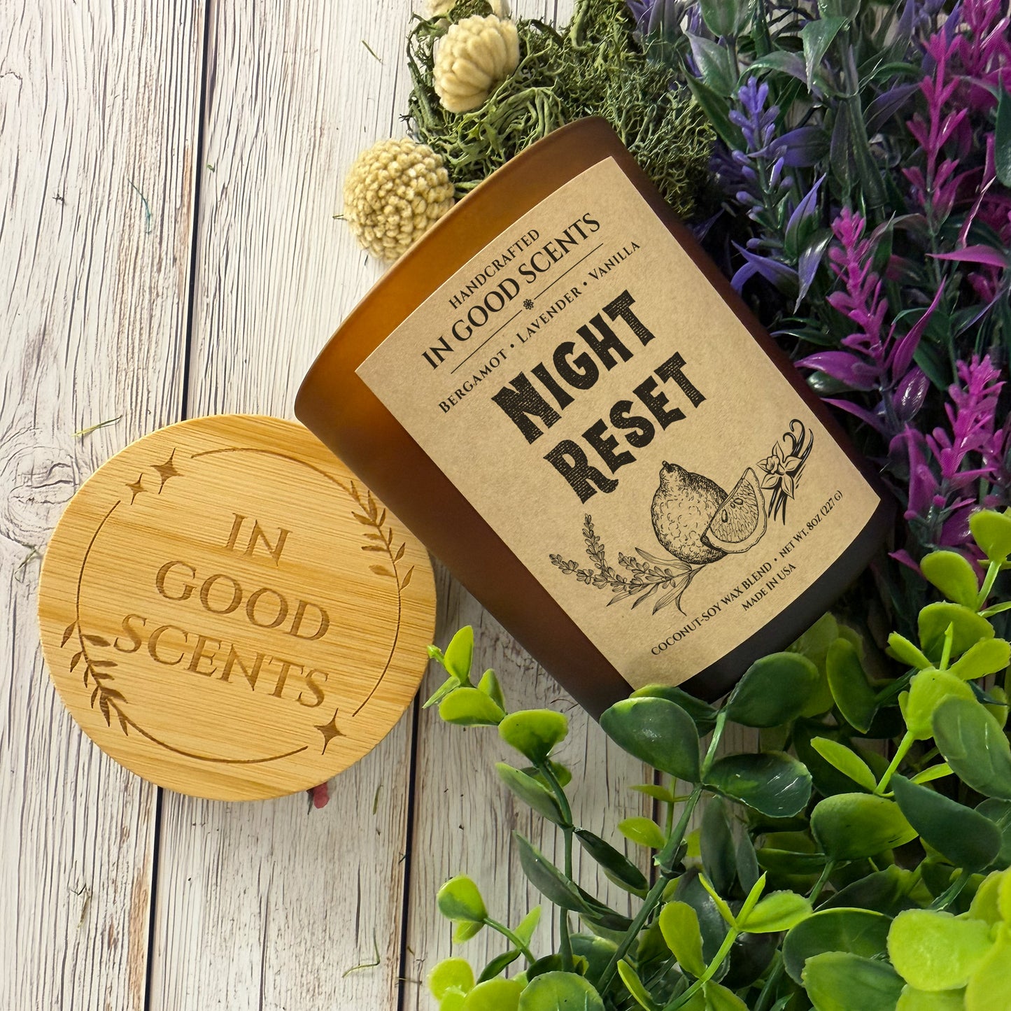 Top-down styled Night Reset 8oz candle with bamboo lid, greenery and purple florals on white wood surface (In Good Scents).