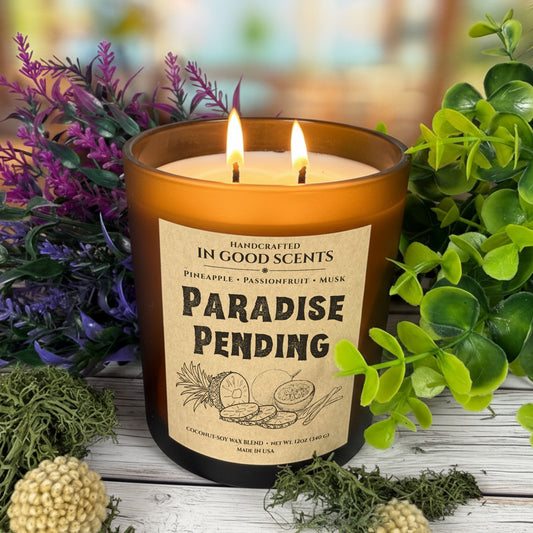 Lit ‘Paradise Pending’ 12oz amber jar candle with two flames, surrounded by greenery and floral decor.