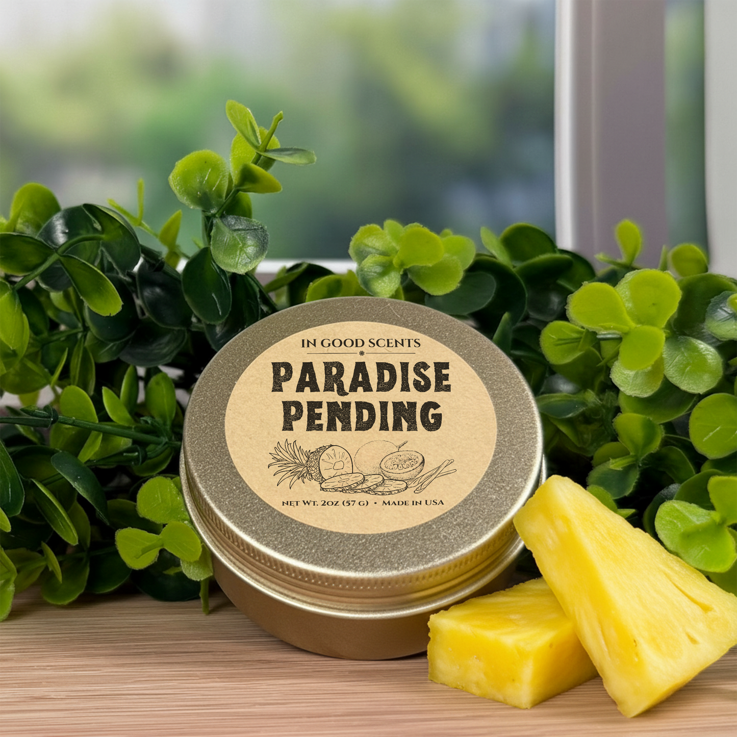 In Good Scents Paradise Pending 2oz travel tin candle on wood surface with greenery and pineapple pieces.