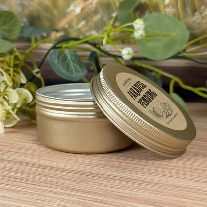 Closed Paradise Pending 2oz travel tin candle with lid label visible on tabletop with soft greenery and flowers.