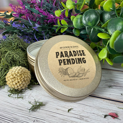 Paradise Pending 2oz travel tin candle with lid resting open beside tin on wood surface with greenery.