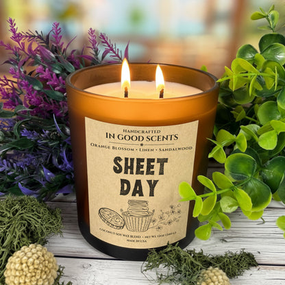 Lit In Good Scents Sheet Day 12oz amber jar candle with two flames, surrounded by greenery on rustic wood background.