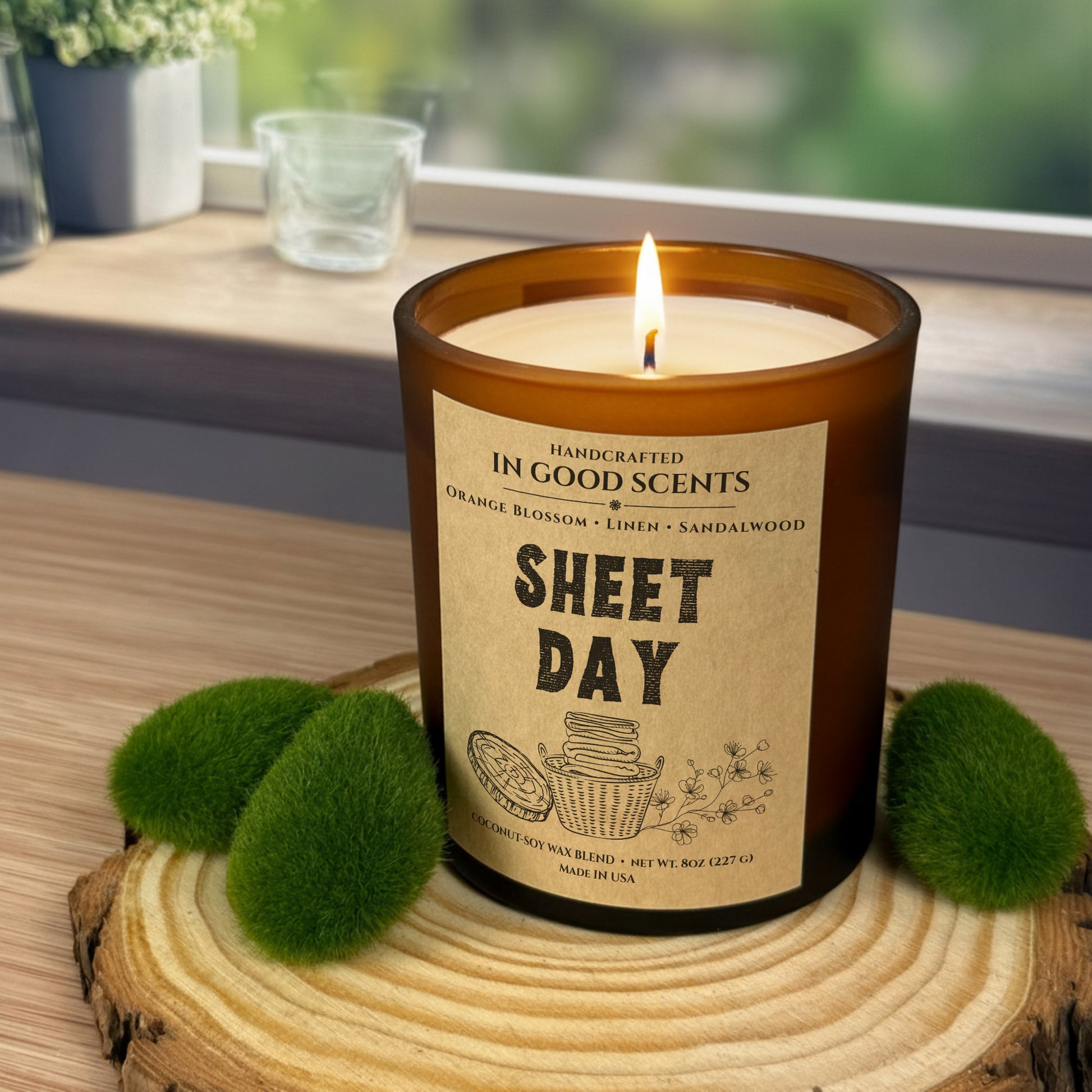 Lit Sheet Day 8oz candle in an amber jar on a wood slice by a window, with soft green moss accents and cozy natural light.