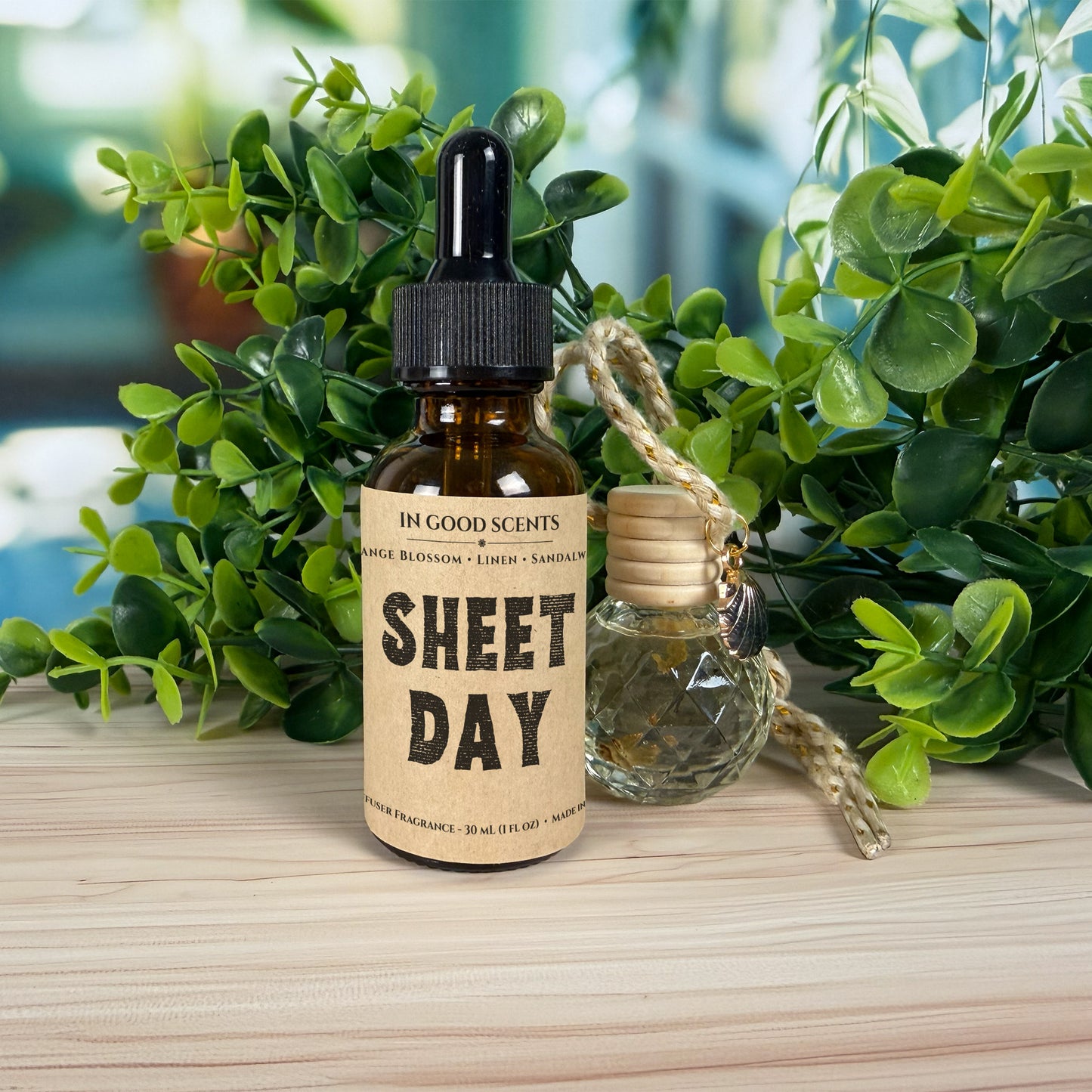 Bottle of 'Sheet Day' diffuser refill with a dropper and wooden cap on a wooden surface with green plants in the background.
