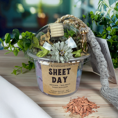 Sheet Day Drive & Unwind gift set cup with hanging car diffuser, wax melts, and gray tassel keychain, styled with greenery on a wooden surface.