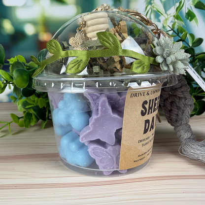 Close-up of Sheet Day Drive & Unwind gift set showing purple and blue wax melts inside a clear cup with decorative ribbon and flower.
